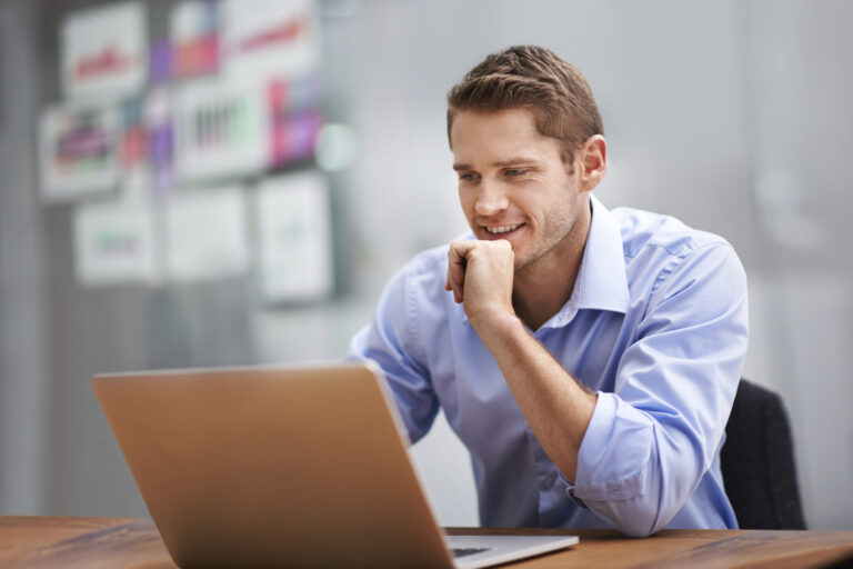 Businessman sitting at desk with hand on chin smiling looking into a laptop.