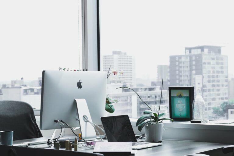 Office skyline and desk with desktop computer, empty chair, mug, plant, and laptop computer.