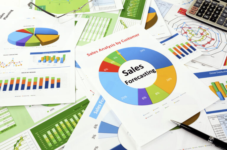 A pile of white papers roughly stacked on a table. They all have colorful graphs, charts, and tables on them. The most prominent one in the center says, "sales forecasting."