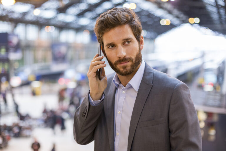 A bearded white man talks on the phone. The background overlooks a crowded office lobby or airport.