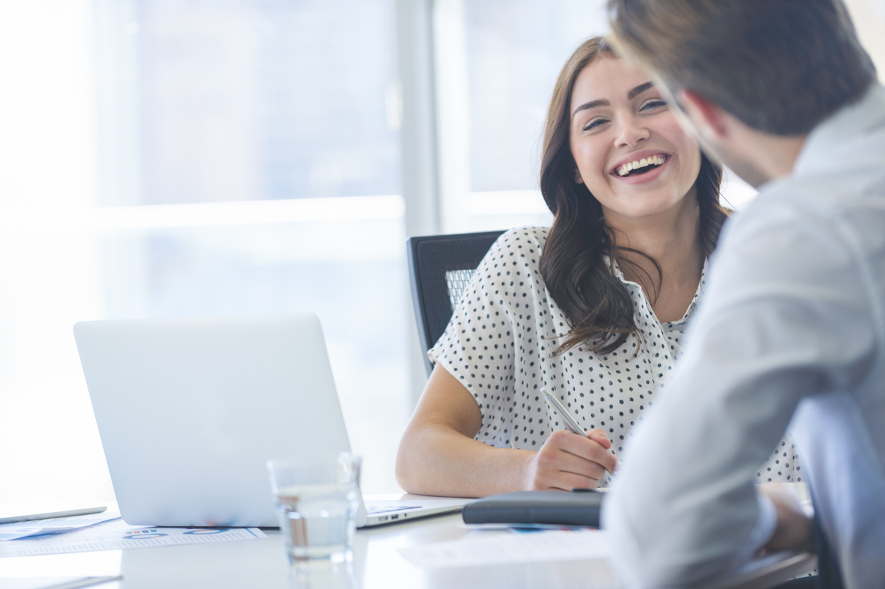 A woman faces the camera as she smiles and appears to be laughing. Across from her is a man who is facing away from the camera. She appears to be taking notes as she has a pen in her right hand. There is a laptop in front of the woman. They appear to be having a meeting.