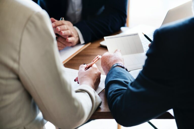 Three professional dressed people sit at a conference table. One of them appears to be taking notes.