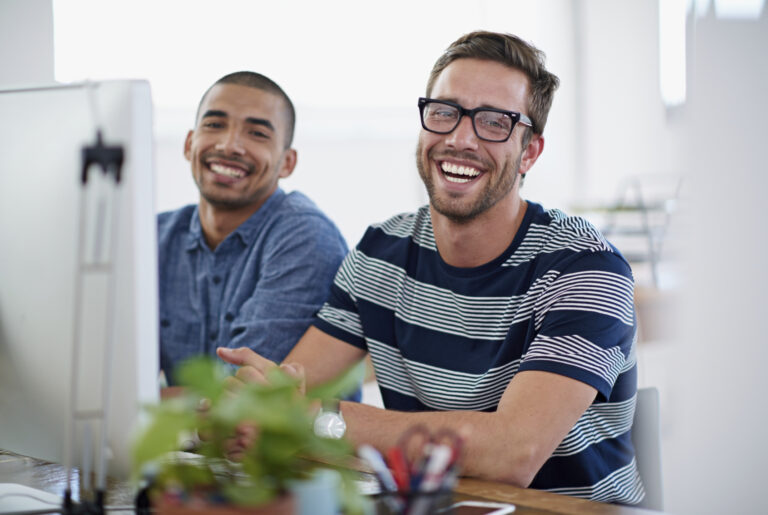 Two casually dressed young bearded men sitting at desk smiling at the camera. The have large desktop computers in front of them.