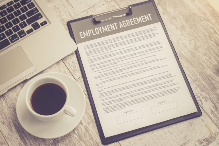In sepia tone, a portion of a laptop is visible next to a cup of coffee and an employment agreement that has yet to be signed.