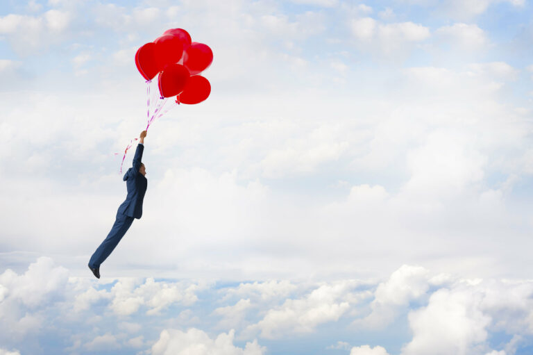 A man flies through sky as he holds on to several red balloons.