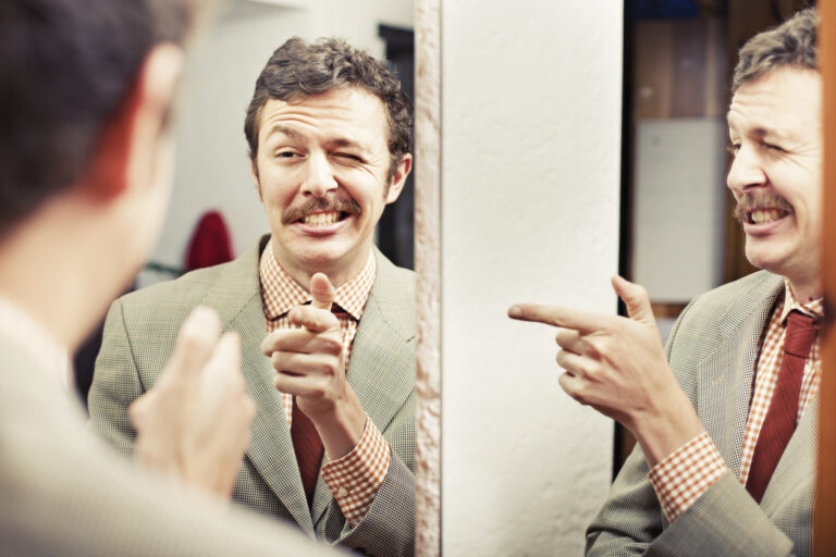 A white man with a mustache is pointing at himself in a mirror and giving himself a cheesy smile. There is a an adjacent mirror that is capturing his reflection from the side too so it looks like there are two of him doing this gesture.