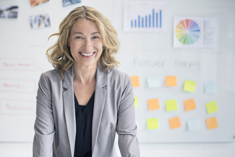 A woman with medium length blond hair smiles at the camera as she leans on something in front of her. There is a white poster behind her labeled "future focus." On the poster is multicolored sticky notes indicating a brainstorming session has occurred.