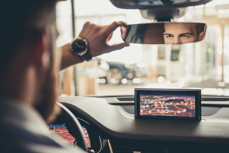 A bearded white man looks through his rear view mirror in a car. He has a map open on his dashboard it what looks like a very high tech car.
