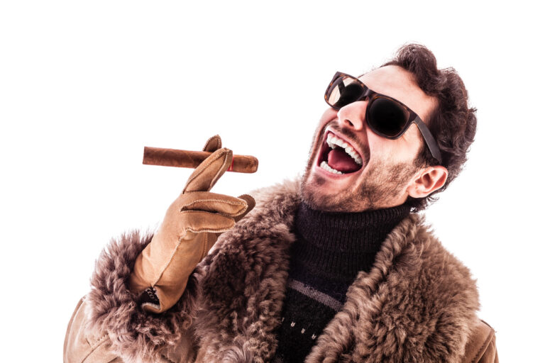 A bearded white man wears sunglasses, a fancy fur coat with leather gloves. He is holding an unlit cigar his right hand fingers and he is smiling in a laughing way. He is leaning back a bit.