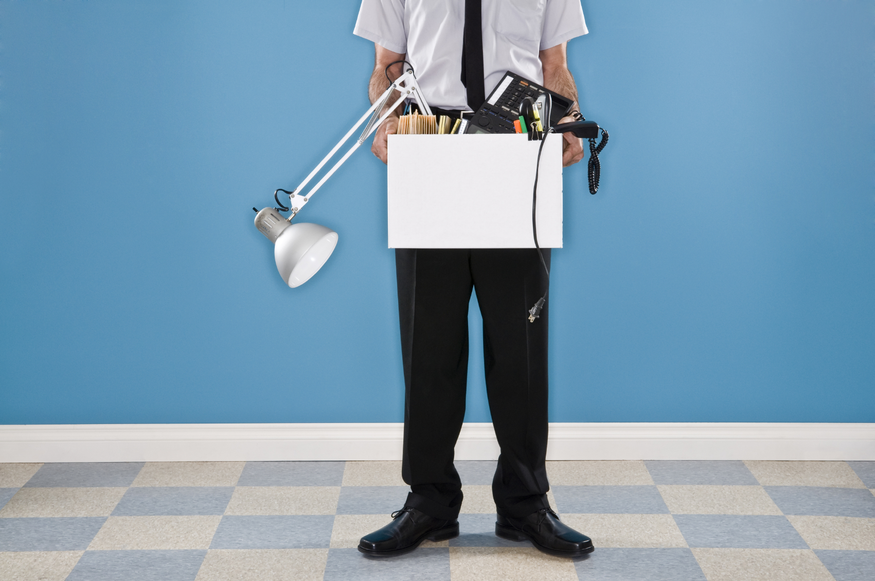A photo of a man from his torso down. He is holding a white box with all his office belongings. He has a short sleeve white shirt and black tie on. His shirt is tucked into black pants.
