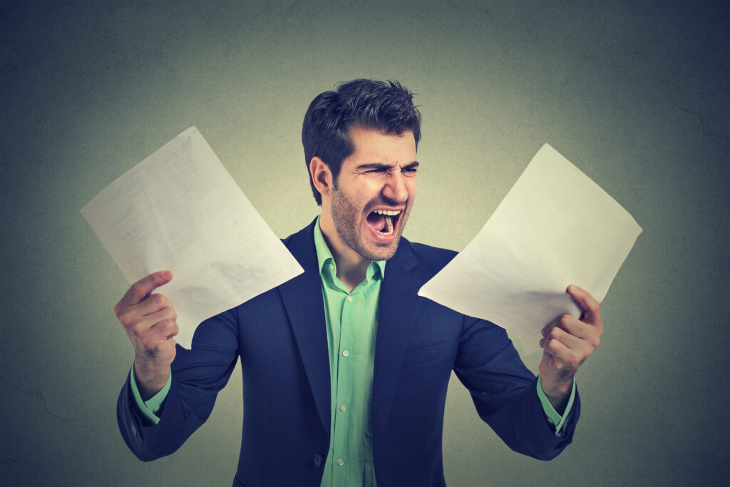 A bearded white man wearing a blue sport coat and green shirt is hold two pieces of paper; one in each hand. He is looking at the one in his left hand and he appears to be shouting.