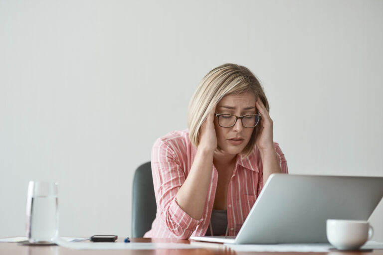 A woman appears to be stressed out as she works in front of her laptop. She is rubbing her temples and has her eyes closed.