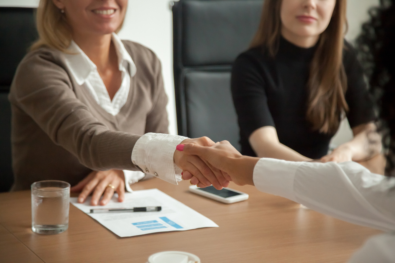 Three women in a conference room. Two of the women are reaching across the table to shake hands.