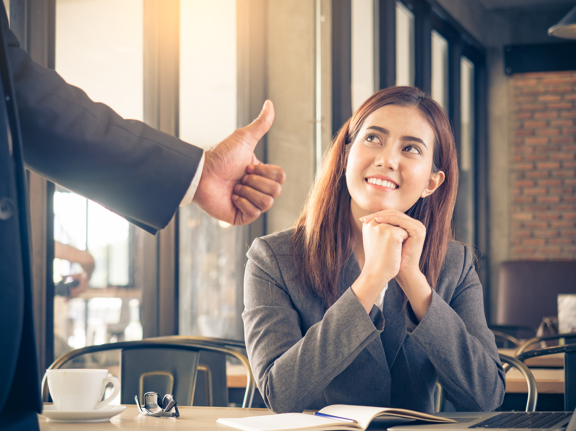 A woman is sitting in a coffee shop with a planner open in front of her. A man gives her a thumbs up.