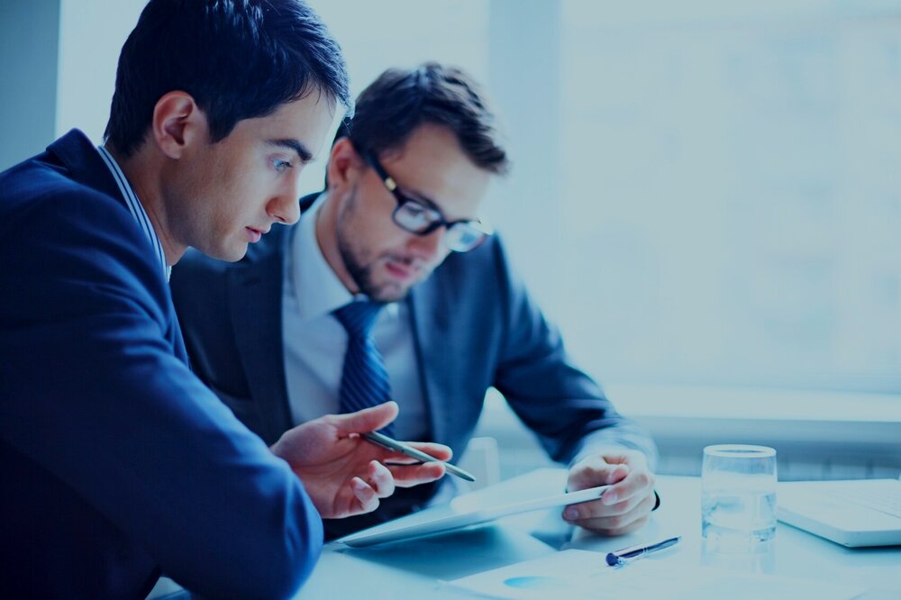 Two men sitting at a desk with water discussing sales hiring.