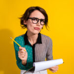 Photo of lovely minded woman wear stylish clothes hold paper list isolated on yellow color background. She's trying to decide which offer to sign.