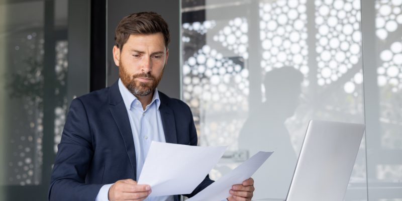 Salesperson in business suit sits in the office at a desk with a laptop and works with documents in his hands